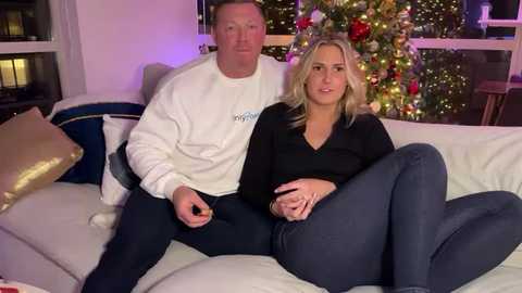 Media: A video of a blonde woman in a black shirt and blue jeans, seated on a white couch beside a man in a white sweater, with a lit Christmas tree in the background.