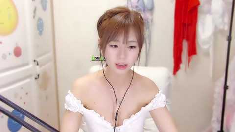 Media: Video of an Asian woman with light skin, brown hair in a bun, wearing a white off-shoulder dress, listening to earphones, in a cluttered, brightly lit room with colorful decor and clothes hanging.