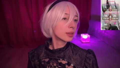 Media: Video of a young woman with short, platinum blonde hair and pale skin, dressed in a black floral patterned dress, standing against a dark red curtain backdrop. A virtual reality headset with a glowing interface is visible.