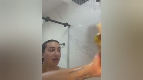 Video of a young woman with dark hair, medium skin tone, and a slim build, showering in a modern bathroom with white marble walls and black metal fixtures. She appears to be applying soap to her body.