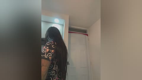 Media: Video of a person with long black hair, wearing a black shirt with a white pattern, standing in a dimly lit bathroom with a white door and shower curtain.