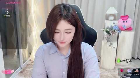 Media: Video of a fair-skinned Asian woman with long brown hair, wearing a light blue button-up shirt, sitting in a black gaming chair, with a pink Kirby plush toy and a bouquet on a side table in the background.