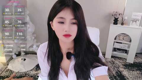 Media: Video of an East Asian woman with long black hair, fair skin, and red lipstick, wearing a white blouse, sitting in a modern, minimalist bedroom with white decor and a patterned rug.