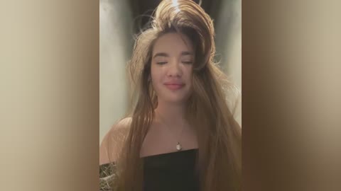 Media: Video of a young woman with long, voluminous brown hair styled high in a messy updo, wearing a black strapless dress, smiling with eyes closed, in a dimly lit, beige-toned background.