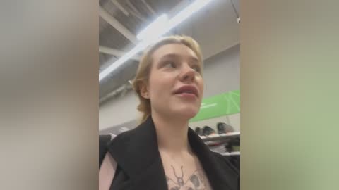 Media: Video of a fair-skinned woman with blonde hair, wearing a black jacket, standing indoors with fluorescent lights overhead. She has a tattoo of antlers on her chest. Background shows a green wall and shelves with various objects.