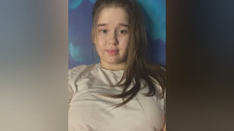 Media: Video of a young Caucasian woman with long brown hair, wearing a white t-shirt, against a blue abstract background. She has a neutral expression.