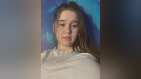 Media: Video of a young, fair-skinned girl with long, light brown hair, wearing a white shirt, against a blue background. She has a neutral expression and is centered in the frame.