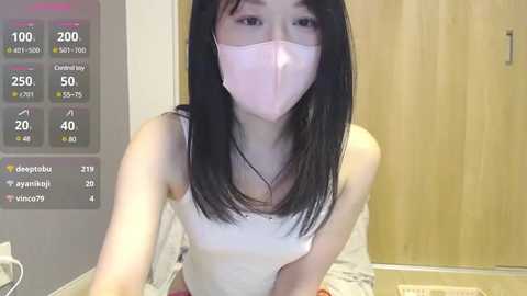 Media: Video of an Asian woman with long black hair, wearing a white tank top and pink face mask, in a bathroom with a wooden door.