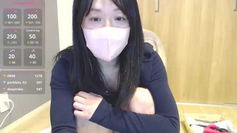 Media: A video of an Asian woman with long black hair and a pink face mask, wearing a navy blue long-sleeve top, sitting on a bed with a wooden headboard.