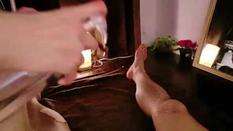 Media: A video showing a massage therapist massaging a client's legs on a dark wooden table. The therapist's hand is covered in oil, while the client's legs are visible, oiled and relaxed. A lit candle and a vase of flowers are on the table's edge.