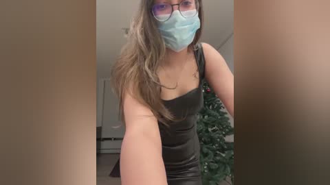 Media: A video of a young woman with long, brown hair, wearing a blue mask, black glasses, and a black tank top, standing in a room with a Christmas tree in the background.