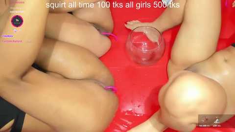 Media: Video of a group of nude women with varying skin tones, positioned on a red inflatable bed. One woman is pouring liquid from a glass onto another's genitals.