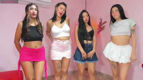 Media: Video of four Latina women in skimpy, revealing outfits, with pink and black tops, high-waisted shorts, and a white skirt, posing in a pink room with a red chair and wall art.