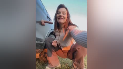 Media: A video captures a woman with long brown hair, wearing a white crop top and gray pants, squatting beside a blue car on a grassy field. She has a smile, and the image is framed by a vertical split.