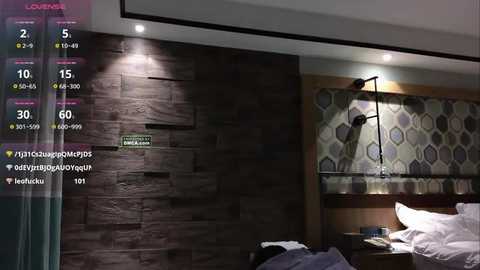 Video of a modern, dimly lit bedroom with a dark wood-textured wall, geometric-patterned wallpaper, and a white bed with rumpled sheets. A black desk lamp hangs from the ceiling.