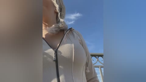 Media: A video of a woman's torso in a beige zip-up hoodie, with blonde hair and a black necklace, standing outdoors against a blue sky backdrop.