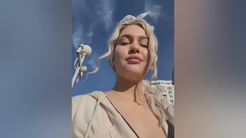 Video of a blonde woman with closed eyes, wearing a beige jacket and black top, with a white orb hovering near her head against a clear blue sky.