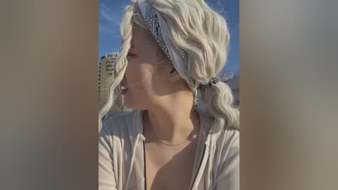 Media: Video of a fair-skinned woman with wavy blonde hair, wearing a white headband and open beige cardigan, standing outdoors with a clear blue sky and distant city buildings in the background.