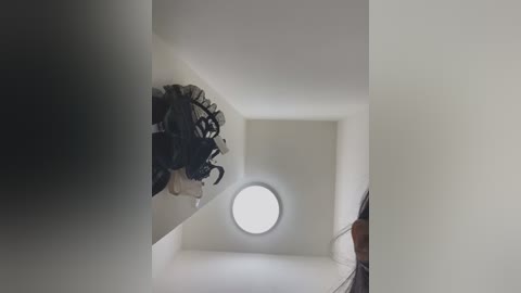 Media: Video of a minimalist, white-walled hallway with a black, abstract, three-dimensional sculpture mounted on the wall. A round, bright window at the end of the hallway emits soft light, creating a serene, modern atmosphere.