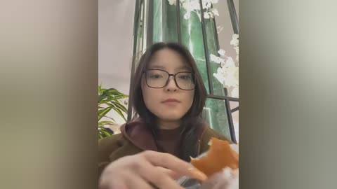 Media: Video of a young woman with straight black hair, wearing glasses, a brown coat, and a maroon scarf, holding an orange, against a background of green curtains and white orchids.