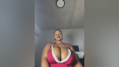 Media: Video of a plus-size Black woman with large breasts, wearing a red halter top with white lace trim, standing in a dimly lit, sparsely furnished room with a ceiling light.