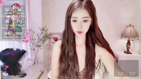 Media: Video of a young East Asian woman with long, straight brown hair, fair skin, and wearing a leopard-print bra. Background includes a white lamp, floral decor, and a pink cushion.