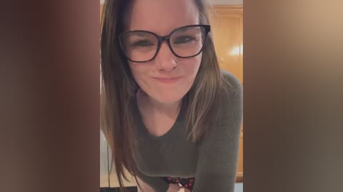 Media: A close-up video of a Caucasian woman with long brown hair, wearing black-rimmed glasses, smiling slightly, and a green sweater, indoors with wooden furniture and a blurred background.