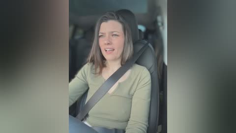 Video of a smiling white woman with shoulder-length brown hair, wearing a green sweater, seated in a car with a seatbelt across her chest.