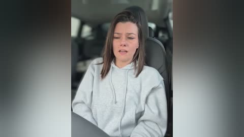 Media: Video of a young woman with shoulder-length brown hair, wearing a light gray hoodie, sitting in a car with the windows blurred, appearing tired and relaxed.