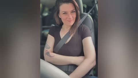 Media: Video of a smiling, fair-skinned woman with long brown hair, wearing a black shirt, seated in a car, with a seatbelt across her chest, and a tattoo on her left arm.