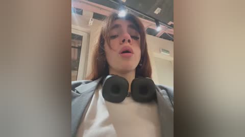 Media: Video of a young person with long, straight brown hair, wearing a grey jacket over a white shirt, and large black headphones. The background features a dimly lit room with exposed ceiling pipes and a door.