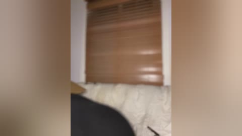 Media: A blurry video shows a bed with beige walls and a brown wooden shutter partially open, casting shadows. The bedding is white with a black blanket. The image appears to be taken indoors.