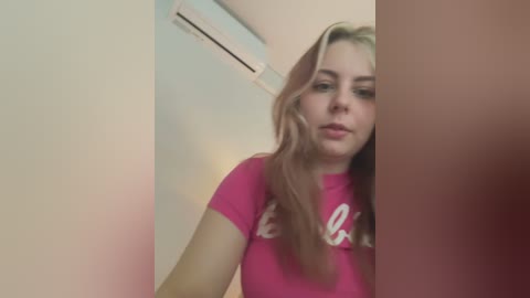 Media: Video of a young Caucasian woman with long, light brown hair, wearing a pink T-shirt with white text, standing indoors next to a beige wall with an air conditioner.