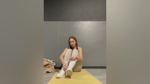 Media: Video of a slender, light-skinned woman with long, straight brown hair, sitting on a yellow yoga mat in a minimalist, gray-walled room, wearing beige leggings and white sneakers, adjusting her socks.