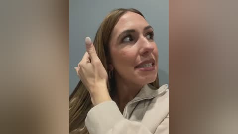 Media: Video of a Caucasian woman with long, straight brown hair, wearing a beige hoodie, adjusting her hair with her right hand. Background is a blurred, light blue wall.