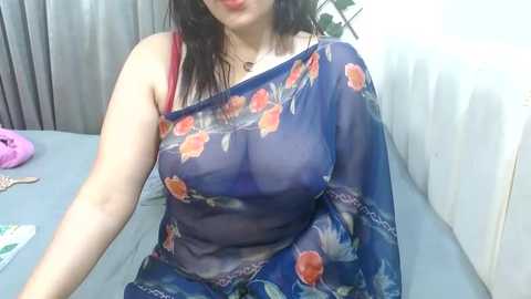 Media: Video of a woman with fair skin, dark hair, wearing a sheer blue floral blouse revealing her large breasts and red bra. She sits on a white couch, surrounded by purple and pink clothes.