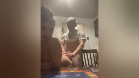 Media: Video of a naked woman on a table, being penetrated by a man in a grey t-shirt and cap, in a dimly lit, sparsely furnished room with a TV.