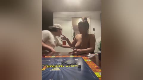 Media: Video of two topless women in a dimly-lit room, one wearing a white hat, the other a bun. A Monopoly board and a glass of red liquid are on the table.