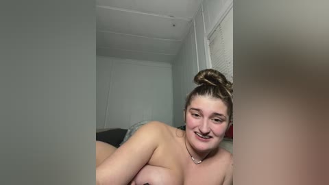 Media: A video of a smiling, nude, plus-sized woman with light skin, brown hair in a bun, and medium-sized breasts lying on a bed in a sparsely decorated room with white walls and a window.