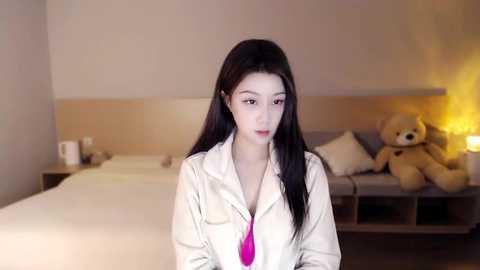 Media: A video of a young Asian woman with long black hair, wearing a white button-up shirt with a pink tie, standing in a modern bedroom with a beige headboard, plush teddy bear, and warm lighting.