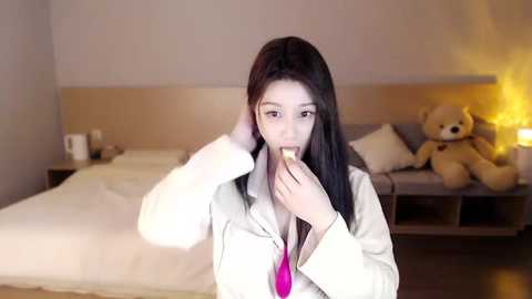 Video of a young woman with long dark hair, wearing a white shirt and pink tie, eating a sandwich in a modern bedroom with beige walls, a wooden bed, and a teddy bear.