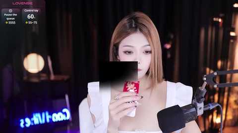 Media: Video of an Asian woman with shoulder-length, straight, light brown hair, wearing a white top, holding a red phone, speaking into a microphone, on a dimly lit stage.