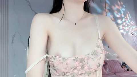 Media: Video of a light-skinned woman with long dark hair, wearing a revealing, lace-trimmed, pastel-colored camisole, exposing her small breasts. Background features a soft-focus, light-colored wall with a string of lights casting a warm glow.