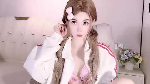 Media: Video of a young Asian woman with light skin, long blonde hair, and a white hair clip, wearing a white jacket and a pink lace bra, sitting in a white room with a flower vase on a white table.