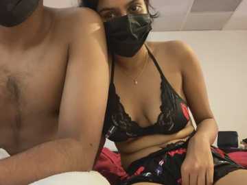 Media: Video of a topless man with a mask, sitting next to a woman in black lingerie and a mask, in a dimly-lit room with white walls.