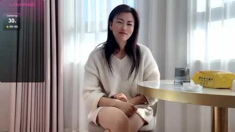 Media: Video of a young Asian woman with long black hair, sitting in a cozy room with light curtains and a round table. She wears a white sweater and beige skirt.