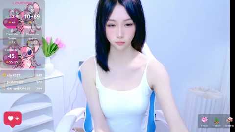 Media: A video of a young Asian woman with straight black hair, wearing a white tank top, sitting in a white room. Digital overlays show game notifications and a live stream interface.