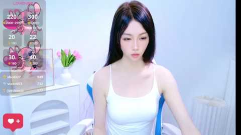 Media: Video of a young Asian woman with long black hair, wearing a white tank top, sitting on a bed in a minimalist bedroom.