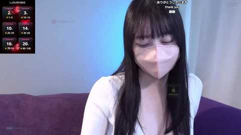 Media: Video of a young Asian woman with long black hair and fair skin, wearing a white mask and a white top, sitting on a purple couch, with a weather app showing rain in the background.