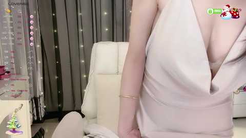 Media: A video captures a woman in a revealing, sleeveless white dress with a deep V-neckline, revealing ample cleavage, standing indoors next to a white leather chair.